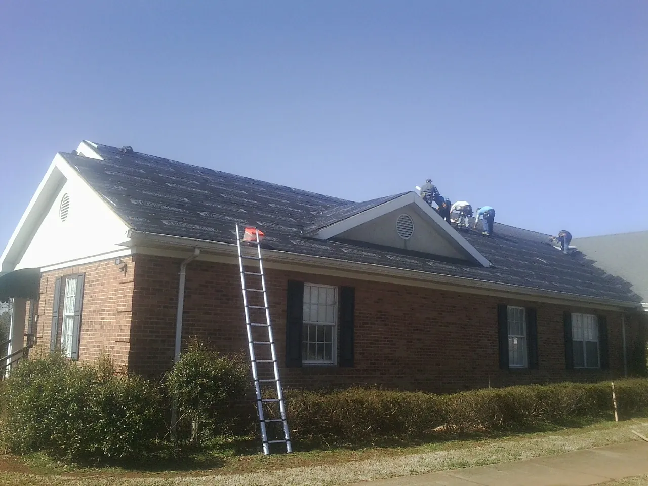 Crew tearing shingles off a brick ranch roof with a ladder set — Metal Roof Repair work by our crew in Rickman