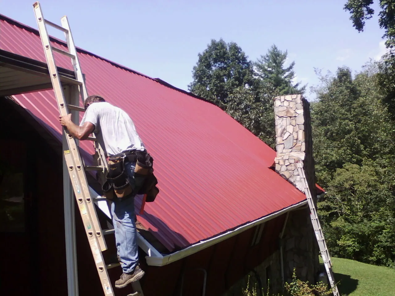 Roofer on a ladder beside a red metal roof and stone chimney — Soffit & Fascia Repair by Roofing Brothers in Rickman